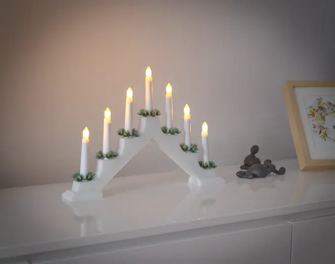 Hvid elektrisk lysestage med syv LED-lys, hver pyntet med små grønne grangrene, arrangeret i en buet form. Velegnet til jule- eller festdekoration.