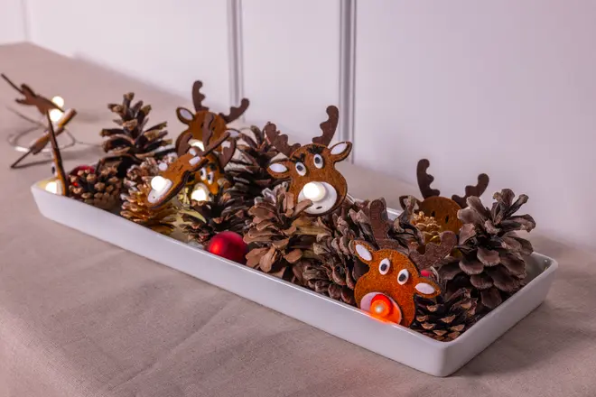 Rektangulær hvid bakke med kogler, røde kugler og rensdyrformede fyrfadsstager. Rensdyrene har brune ansigter, gevir og lysende næser, hvilket skaber et festligt julearrangement.