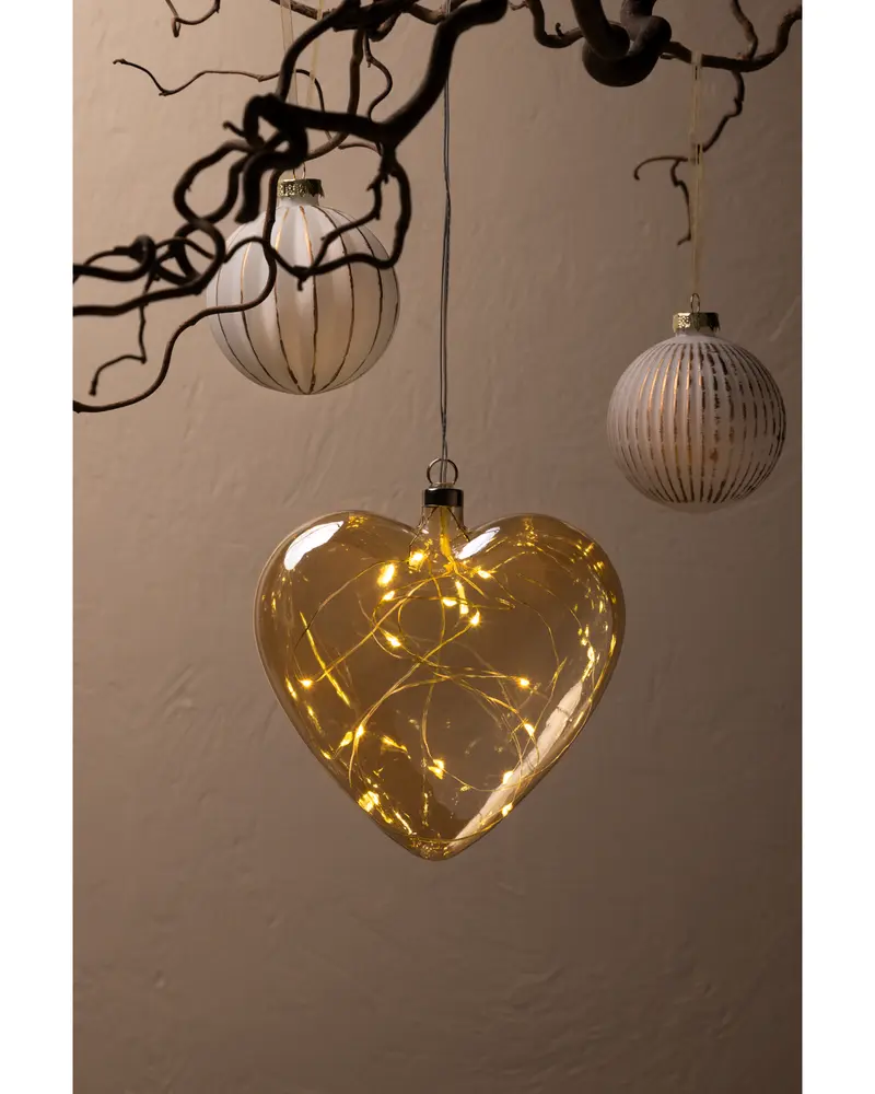 Hjärformad glasprydnad med varma LED-ljusslingor inuti, hängande från en gren. Två runda, randiga glaskulor hänger också nära. Perfekt för festlig eller romantisk dekoration.