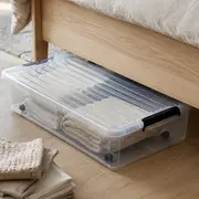 Gennemsigtig under-seng opbevaringskasse med låg og sidehåndtag, fyldt med foldede håndklæder og tøj.