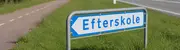 Blåt og hvidt vejskilt med teksten 'Efterskole' og en venstrepil, monteret på metalstænger, placeret på græs ved siden af vej og cykelsti.