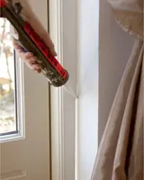 En hand applicerar vit fogmassa med fogpistol längs kanten mellan fönsterkarm och vägg för tätning.