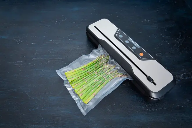 Vakuumpakker, der forsegler en pose med friske asparges. Elegant sølv og sort design med kontrolknapper på toppen. Perfekt til opbevaring af mad.