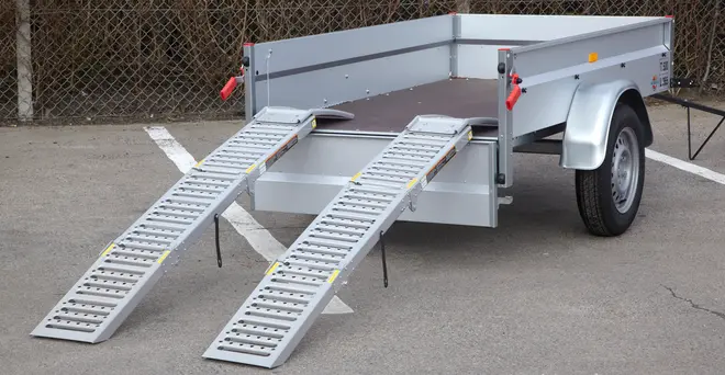 Enkeltakslet trailer med metalsider, åbent lad og to aftagelige læsseramper. Robust hjul, bagreflekser og rummeligt lad til transport af udstyr eller varer.