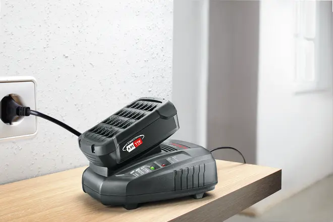 Sort batterioplader med tilsluttet 14,4V lithium-ion batteri, tilsluttet en stikkontakt og placeret på en træoverflade. LED-indikatorer ses på opladeren.