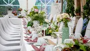 Elegant banketbord dækket med hvide duge, blomsterdekorationer, hvide stole, glas, tallerkener og foldede servietter, ideelt til bryllupper eller formelle arrangementer i pyntet lokale.