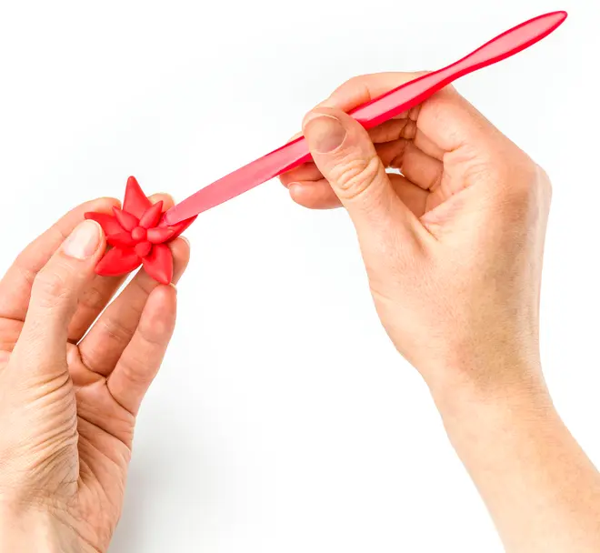 Rødt plastikmodelleringsværktøj med spids ende, bruges til at forme en rød blomst af fondant eller ler, holdt i to hænder mod hvid baggrund.