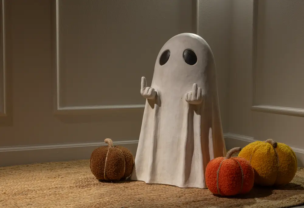 A decorative figure resembling a ghost with a smooth white sheet-like body and two large black oval eyes. The ghost has its arms raised, with both hands making a playful gesture by extending the middle fingers. Surrounding the ghost are three plush pumpkins in brown, orange, and yellow colors, each with a textured surface and a fabric stem. The scene is set indoors on a woven rug against a paneled wall, creating a cozy and festive atmosphere.