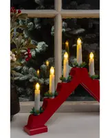 A decorative candle holder with five electric candles arranged in a stepped, triangular formation. The base is painted red, and each candle is white with a warm yellow flame-shaped bulb. Small green artificial pine sprigs with red berries are placed at the base of each candle for a festive touch. The candle holder is displayed on a windowsill with a snowy outdoor scene visible through the window.