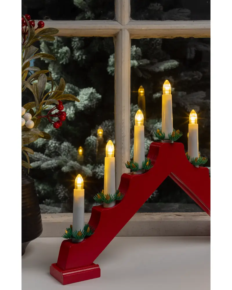 A decorative candle holder with five electric candles arranged in a stepped, triangular formation. The base is painted red, and each candle is white with a warm yellow flame-shaped bulb. Small green artificial pine sprigs with red berries are placed at the base of each candle for a festive touch. The candle holder is displayed on a windowsill with a snowy outdoor scene visible through the window.