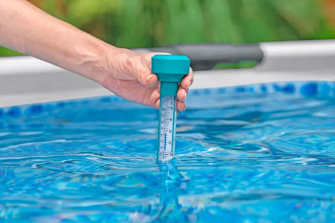 Hånd holder et flydende pooltermometer med tydelig temperaturskala, delvist nedsænket i blåt vand, bruges til præcis måling af pool- eller spavandstemperatur.