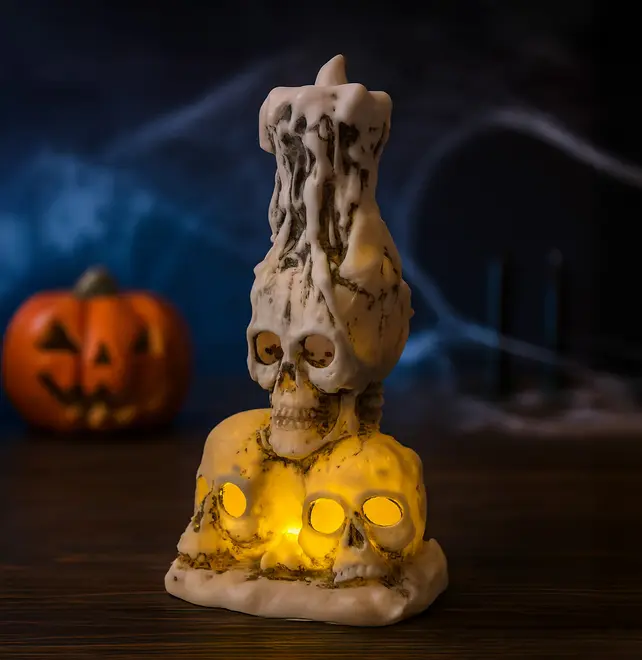 A decorative candle holder designed to resemble three realistic skulls at the base, with hollow eye sockets that emit a warm yellow glow from internal lights. The top features a larger skull with a melted wax effect, giving the appearance of a candle dripping over the skull. The overall finish is aged and weathered, enhancing its spooky, gothic aesthetic.