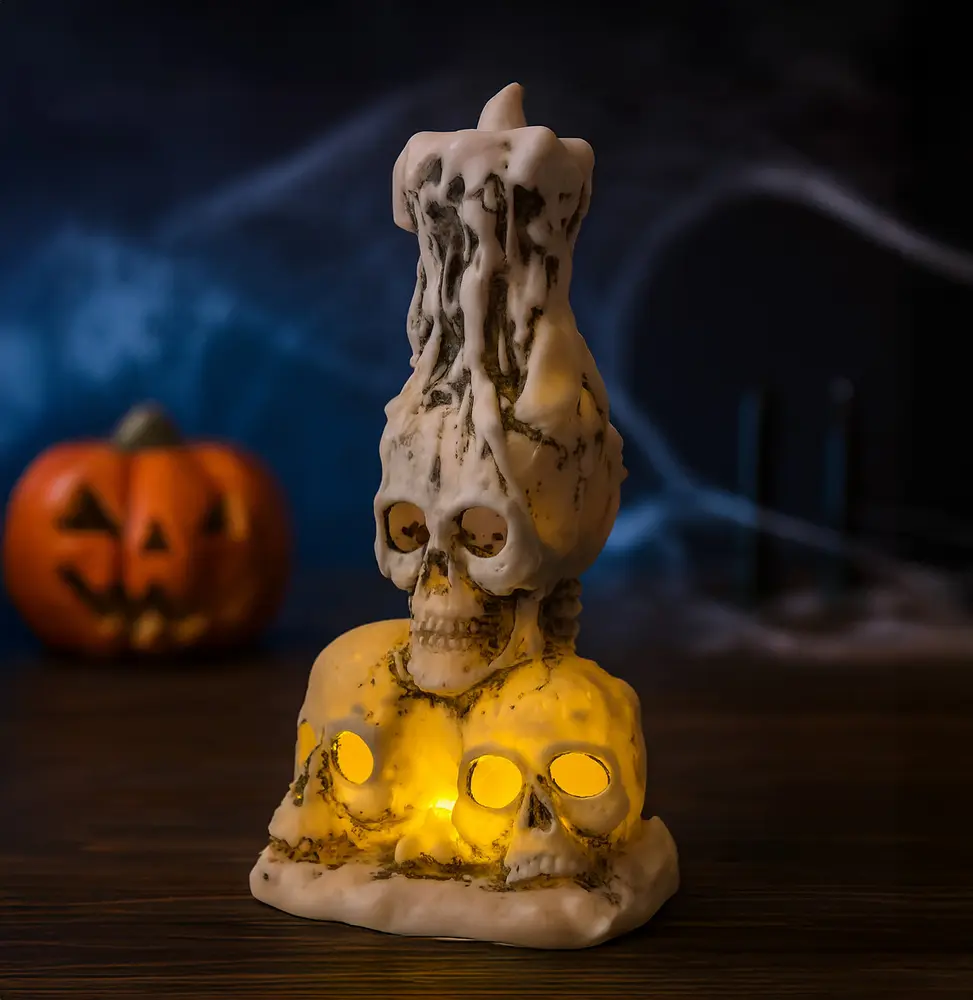 A decorative candle holder designed to resemble three realistic skulls at the base, with hollow eye sockets that emit a warm yellow glow from internal lights. The top features a larger skull with a melted wax effect, giving the appearance of a candle dripping over the skull. The overall finish is aged and weathered, enhancing its spooky, gothic aesthetic.
