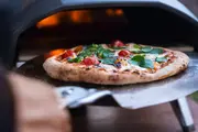 Træfyret pizza med tyk, luftig skorpe, toppet med smeltet ost, cherrytomater og friske basilikumblade, tages ud af en pizzaovn med en metalspade.