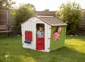 Plastik legehus til børn med brunt tag, hvide vægge, grøn side, rød dør, røde vinduesskodder og blå vindueskarm. Velegnet til udendørs leg i haven.