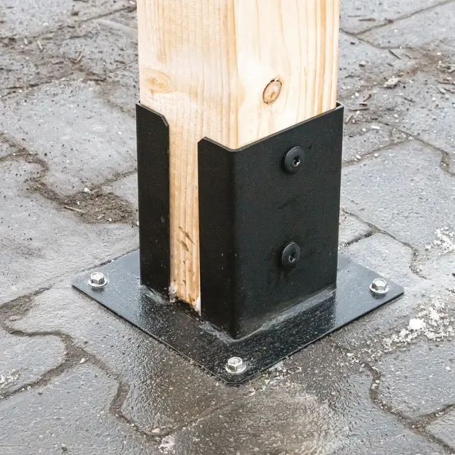 Metal stolpebeslag fastgjort til beton med bolte, holder en lodret træbjælke. Giver stabil støtte til udendørs konstruktioner som hegn, pergolaer eller terrasser.