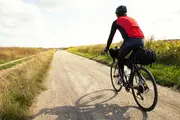 Cyklist iført rød og sort trøje, sort hjelm og tights, cykler på en touringcykel med bagtasker på en grusvej gennem åbent landskab.