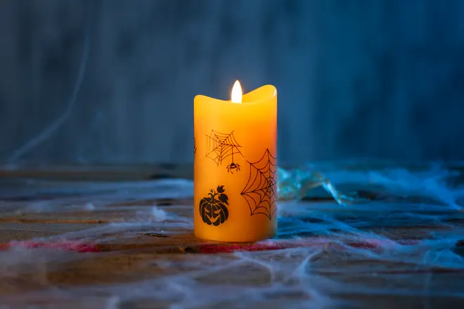 Gult pelarljus med Halloweenmotiv, inklusive spindelnät, en spindel och en pumpa. Ljuset är tänt och står på en träyta med konstgjorda spindelnät runtom.