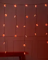 String lights with small, round bulbs emitting a warm orange glow. The lights are arranged in vertical strands hanging from a horizontal wire, creating a curtain effect. The bulbs are evenly spaced along each strand, and the wires are dark, making the glowing bulbs stand out against the dimly lit background.