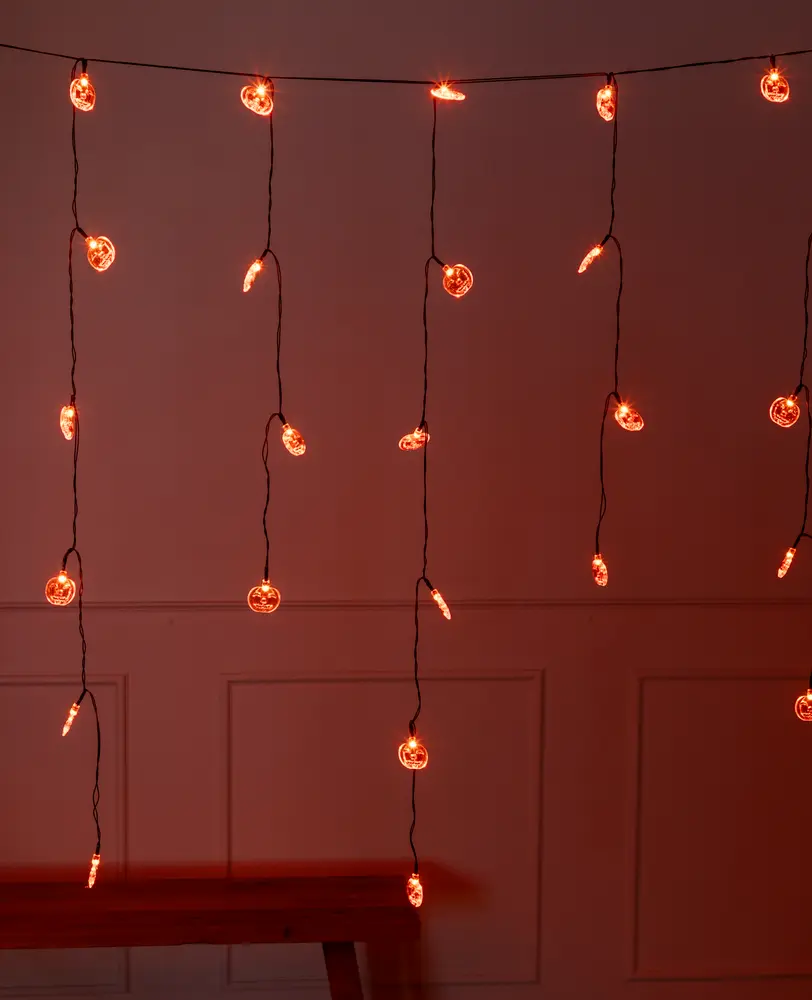 String lights with small, round bulbs emitting a warm orange glow. The lights are arranged in vertical strands hanging from a horizontal wire, creating a curtain effect. The bulbs are evenly spaced along each strand, and the wires are dark, making the glowing bulbs stand out against the dimly lit background.