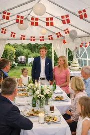 Udendørs festbord under hvidt telt med danske flag, hvide duge, blomsterdekorationer, tallerkener med mad, stearinlys og balloner. Festlig stemning til fejring eller sammenkomst.