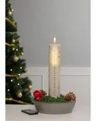 Högt vitt adventsljus med siffror 1-24, dekorerat med snöflingor, placerat i rund skål med mossa, kottar och röd kula. Julgran i bakgrunden.