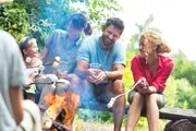 Gruppe mennesker sidder udendørs på en træbænk og rister skumfiduser over et bål, omgivet af grøn natur. Afslappet sommertøj, synlige pinde med skumfiduser og en plastikkop.