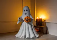 A decorative figure shaped like a friendly ghost with a smooth white sheet-like body, black oval eyes, and a small open mouth. The ghost is holding a carved orange pumpkin with a smiling face. The figure stands on a flat base, and the surface appears to be made of a solid material, possibly resin or plastic. The background features warm lighting and Halloween-themed candles, enhancing the festive atmosphere.