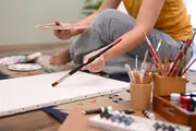 En person forbereder sig på at male på et tomt lærred, omgivet af forskellige pensler, malertuber, en palet og kunstforsyninger, arrangeret på et mønstret tæppe.