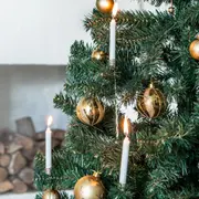 Konstgjord julgran dekorerad med guldfärgade kulor och vita kläm-ljus, några tända, för en festlig och elegant känsla. Gröna grenar ger ett naturtroget utseende.