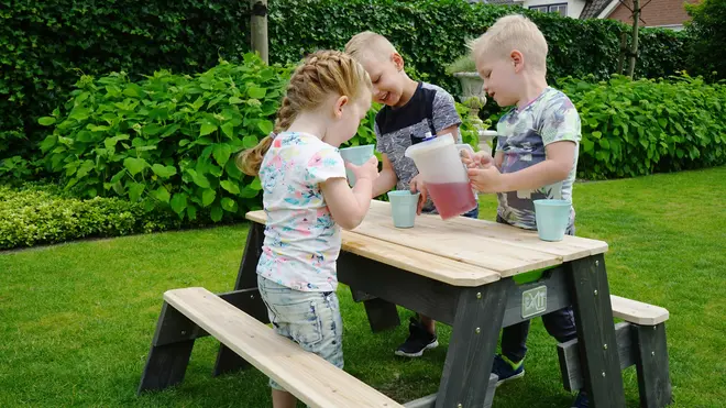 Børnebord i træ med fastmonterede bænke, naturfarvet bordplade og sæder, mørkegrønt stel, velegnet til udendørs brug i have, vist med krus og kande.