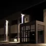 Moderne bygningsindgang med store glasdøre og vinduer, oplyst af udendørs væglamper. Hvid plantekrukke med plante står nær indgangen. Minimalistisk arkitektur.