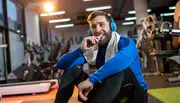 Mand med blå hovedtelefoner og blå langærmet sportstrøje sidder i fitnesscenter med hvidt håndklæde om halsen. Omgivet af træningsudstyr og løbebånd.