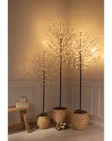 Three decorative artificial trees with slender black branches, each adorned with warm white LED lights at the tips, creating a glowing effect. The trees are placed in woven natural-colored baskets filled with brown filler material. The arrangement includes three different heights for visual interest. The setup is positioned in a softly lit corner, with a wooden bench and wrapped gifts nearby, and gold ornaments on the floor for a festive touch.