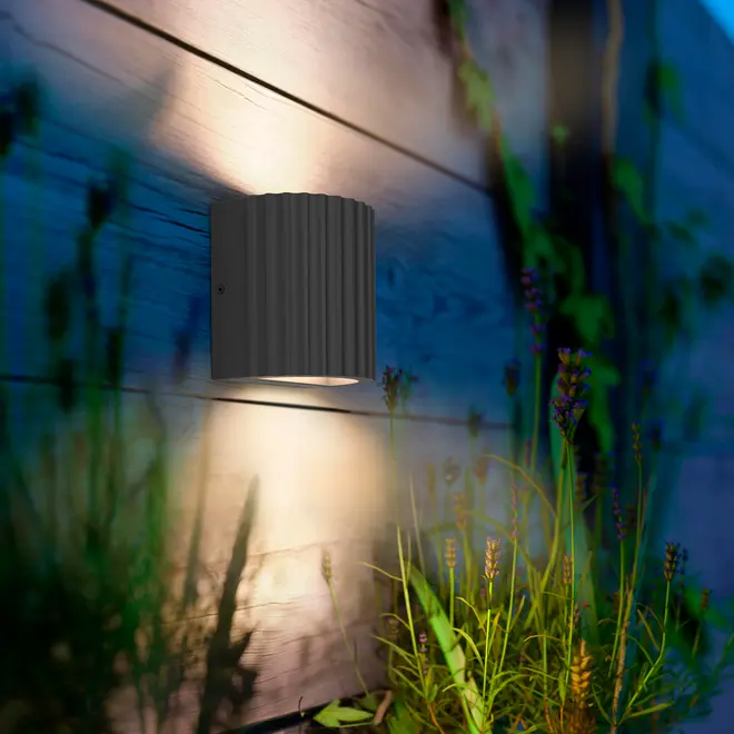 Moderne udendørs væglampe med sort, riflet cylindrisk design. Monteret på trævæg, udsender varm nedadgående belysning, ideel til have eller terrasse.
