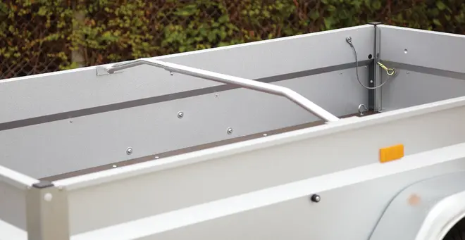 Åben trailerkasse med høje metalsider, tværgående støttebøjle og lysegrå finish. Der er side-reflekser og synlige fastgørelsespunkter indvendigt.