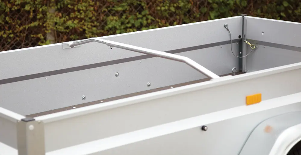 Öppen släpvagn med höga metallsidor, tvärgående stödstång och ljusgrå finish. Sidoreflexer och synliga fästpunkter inuti.