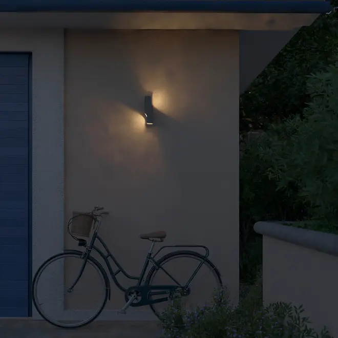 Moderne udendørs væglampe med op- og nedadgående lys, monteret på beige væg. Slankt, rektangulært design i mørk finish, ideel til belysning ved døre eller garager.