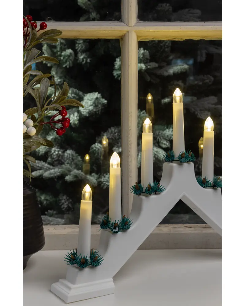 A white, arched candle holder with five electric candles arranged at different heights. Each candle has a warm yellow flame-shaped bulb and is surrounded at the base by small artificial green pine sprigs. The candle holder sits on a white surface in front of a window, with a festive plant featuring red and white berries nearby.