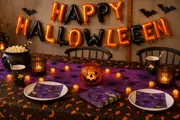 Halloween-borddækning med orange og sorte ballonbogstaver, græskarlygte, flagermusdekorationer, lilla servietter, tallerkener, kopper, popcorn i gryde og lyskæde med små græskar.