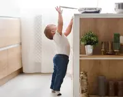 Ett litet barn i vit tröja och blå jeans står på tå och sträcker sig efter en stekpanna på köksbänken, vilket visar potentiella säkerhetsrisker för barn.