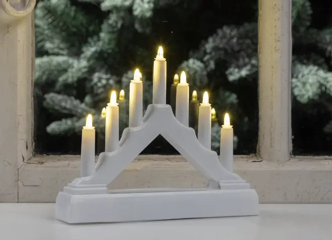 En hvid, trekantet lysestage med syv batteridrevne LED-lys arrangeret i en trappeformet bue. Hvert lys udsender et varmt, gult skær, der ligner ægte flammer. Basen er solid og rektangulær, og det samlede design er enkelt og moderne, velegnet til vindueskarm eller bord.