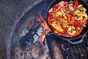 Støbejernspande med grillet oksekød, rød peberfrugt, løg og gul squash, tilberedt over bål. Friske, farverige ingredienser perfekte til udendørs madlavning og camping.
