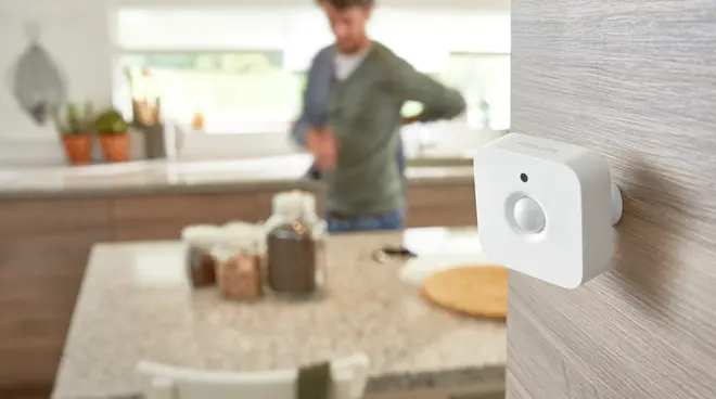 Hvid, firkantet bevægelsessensor med afrundede hjørner, monteret på væg. Har en central sensor og lille indikatorlys. Moderne design, velegnet til smart home-sikkerhed.