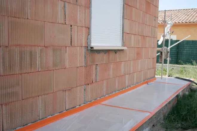 Ufærdig murstensvæg med hvidt vinduesskodde. Jorden er dækket med beskyttende plastfolie fastgjort med orange tape, sandsynligvis til bygge- eller malerarbejde.