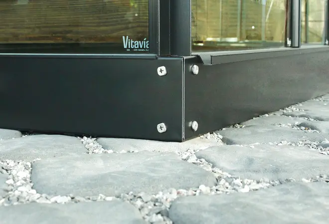 Närbild av svart metallram till växthus med synliga skruvar, monterad på gatsten. Glasrutor sitter ovanpå ramen. Märket 'Vitavia' syns.