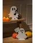 Two white ghost-shaped decorations with black eyes and mouth, each standing on a flat base. One ghost is holding a black bottle with a skull and crossbones design, while the other ghost is holding a rectangular sign that says 'BOO' in black letters. The scene is decorated with small, textured pumpkin ornaments in orange and yellow, and two lit pumpkin-shaped candles, creating a warm, festive Halloween atmosphere.