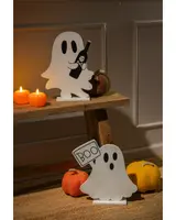 Two white ghost-shaped decorations with black eyes and mouth, each standing on a flat base. One ghost is holding a black bottle with a skull and crossbones design, while the other ghost is holding a rectangular sign that says 'BOO' in black letters. The scene is decorated with small, textured pumpkin ornaments in orange and yellow, and two lit pumpkin-shaped candles, creating a warm, festive Halloween atmosphere.