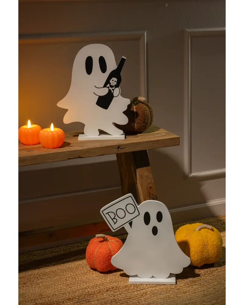Two white ghost-shaped decorations with black eyes and mouth, each standing on a flat base. One ghost is holding a black bottle with a skull and crossbones design, while the other ghost is holding a rectangular sign that says 'BOO' in black letters. The scene is decorated with small, textured pumpkin ornaments in orange and yellow, and two lit pumpkin-shaped candles, creating a warm, festive Halloween atmosphere.