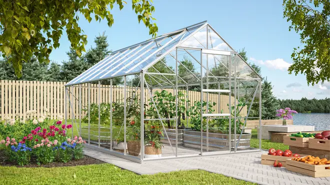 Växthus av klart glas med aluminiumram, sadeltak och dörr. Innehåller olika gröna växter i krukor och bäddar. Placerat på stenlagd yta i trädgård.